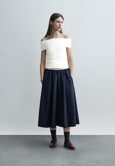 Jeune femme portant un haut blanc à épaules dénudées, une jupe midi bleu marine, des chaussettes foncées et des chaussures bordeaux, debout devant un fond gris clair uni.