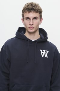 Giovane uomo con capelli ricci che indossa un hoodie blu navy con il logo bianco "VA" sul petto, rivolto in avanti contro uno sfondo bianco semplice.