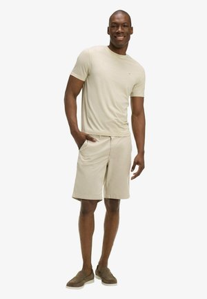 Homme souriant debout avec une main dans la poche, portant un t-shirt beige, un short assorti et des chaussures marron sans lacets sur fond blanc.