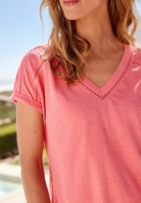 Bluzka z dekoltem w serek, krótki rękaw w intensywnym koralowym kolorze, wykonana z miękkiego materiału z koronkowym wykończeniem na ramionach. Gładka tekstura, luźny krój i podwinięte rękawy.