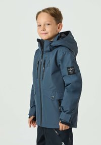 Giacca impermeabile blu marina con cappuccio, dotata di multiple tasche con cerniera, dettagli neri e una toppa con il logo sulla manica.