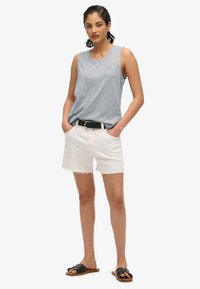 Canotta grigia senza maniche, pantaloncini di denim bianco sfilacciati, cintura nera e sandali scuri, presentati su uno sfondo bianco semplice.