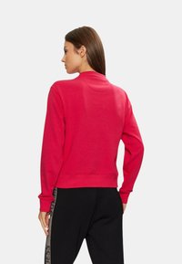 Sudadera roja con cuello redondo, puños y dobladillo de canalé. Vista desde la parte posterior, combinada con pantalones negros con una franja lateral estampada.