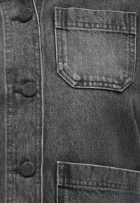 Details van een grijze denimjas met knoopsluiting, een bovenste zak in octagonale vorm en een gestructureerde stof.