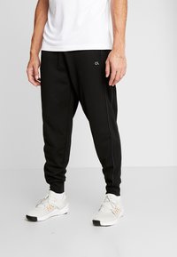 Zwarte trainingsbroek met een taps toelopende pasvorm, voorzien van zijnaad piping en een logo aan de linkerkant, gecombineerd met witte sportschoenen.