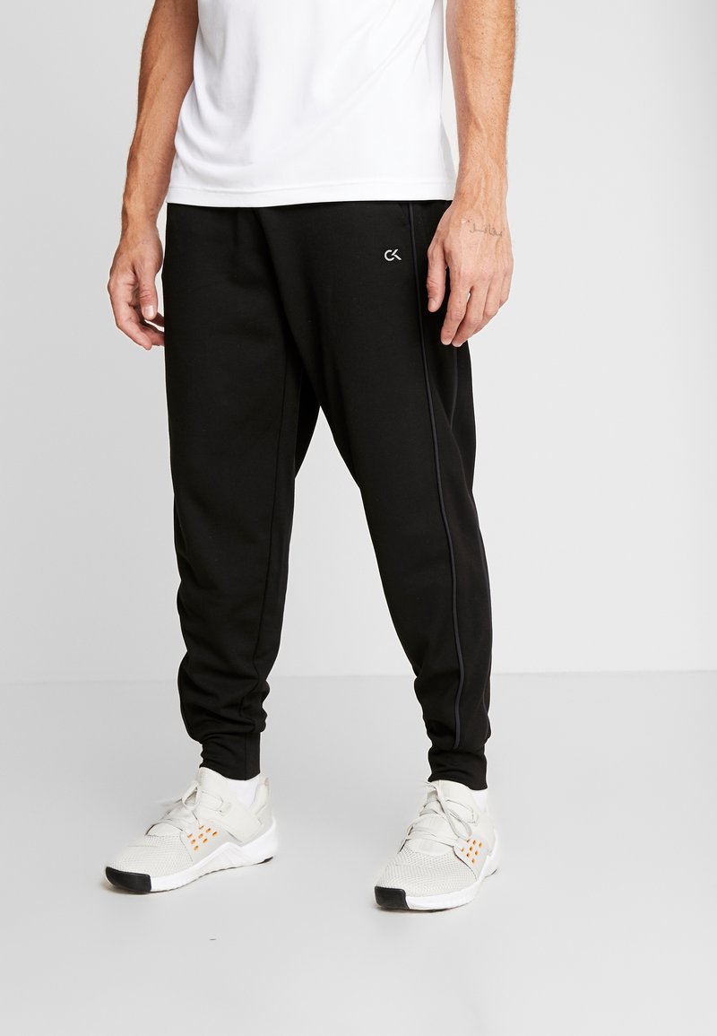 Zwarte trainingsbroek met een taps toelopende pasvorm, voorzien van zijnaad piping en een logo aan de linkerkant, gecombineerd met witte sportschoenen.