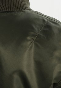 Ciemnozielona kurtka z prążkowanym kołnierzem, gładką fakturą i subtelnym połyskiem, z detalem szwu na ramieniu dla zwiększenia walorów projektowych.