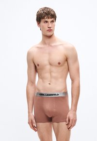Boxer cortos de algodón marrón con una cinturilla gris que muestra el logo "KARL LAGERFELD". Textura suave, diseño ajustado, estilo minimalista.