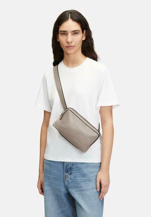 Graue Ledertasche zum Umhängen mit schwarzem Reißverschlussakzent, rechteckige Form, geprägter Markenname, getragen über einem weißen T-Shirt und blauer Jeans.