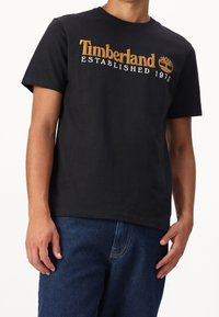 Černé bavlněné tričko s potiskem "Timberland Established 1973" ve zlatých a bílých barvách, s kulatým výstřihem a krátkými rukávy.
