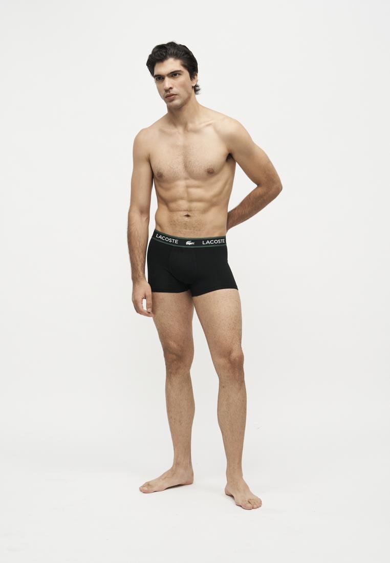Homme torse nu, pieds nus, portant un boxer Lacoste noir sur fond blanc en studio.