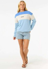 Sudadera color bloqueada en azul y beige, con mangas largas y cuello redondo, que presenta un pequeño logo en el pecho. Combinada con pantalones cortos de mezclilla.
