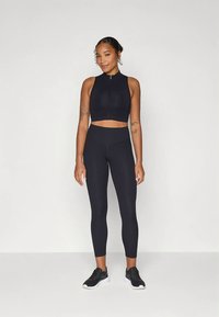 Černý atletický crop top s vysokým límcem a předním zipem, doplněný o černé legíny s vysokým pasem a černé atletické boty. Hladká textura.