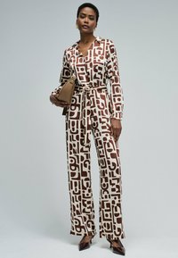 Tuta stampata in crema e marrone con motivi geometrici, scollo con colletto, vita con cintura e design a gamba larga; accessoriata con una borsa color cuoio.