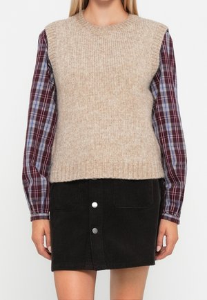 Femme portant un pull sans manches beige en tricot par-dessus une chemise à carreaux grenat et une mini-jupe noire boutonnée, debout devant un fond blanc.