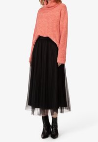 Maglione a collo alto in colore corallo abbinato a una gonna nera in tulle plissettato e stivaletti neri con punta. L'insieme combina diverse trame.