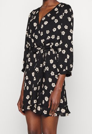 Vestido cruzado negro con estampado floral blanco, mangas tres cuartos y cinturón anudado en la cintura, llevado por una persona con piel oscura.