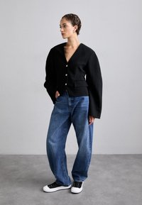 Svart teksturert blazer med dyp V-hals og sølvknapper, kombinert med løstsittende blå jeans og svart-hvite sneakers.