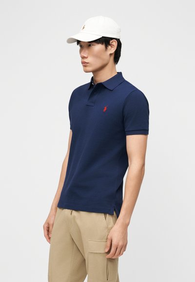 Uomo che indossa una polo blu navy con logo rosso, pantaloni beige con tasca cargo e un cappellino da baseball bianco, in piedi di lato.