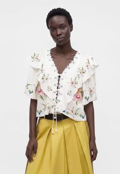 Femme portant un chemisier blanc brodé de fleurs avec volants et noué devant, ainsi qu'une jupe plissée jaune avec ceinture noire sur un fond uni.