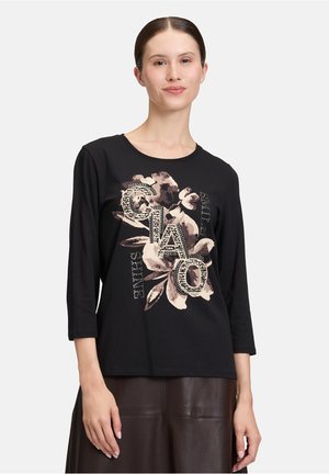 Donna che indossa una maglietta nera a maniche lunghe con un motivo floreale beige e la parola "GIAO" in grandi lettere decorative sulla parte anteriore.