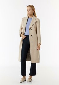 Beżowy wełniany płaszcz z dużymi klapami, dwurzędowy fason i czarne guziki, w zestawie z jasnoniebieskim swetrem i ciemnoniebieskimi dżinsami.