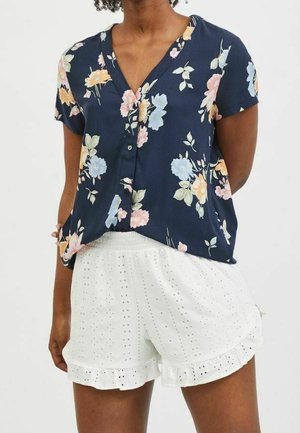Woman wearing a navy blue, short-sleeve floral blouse paired with white eyelet lace shorts with ruffled hems.