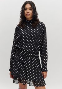 Schwarzes Kleid mit Tupfen, hochgeschlossenem Hals, langen Ärmeln, ausgestellt, zweilagigem Rock und gerafftem Taillenbereich. Hergestellt aus einem leichten, durchsichtigen Stoff.