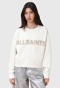 Felpa crema con "ALLSAINTS" in lettere metalliche testurizzate. Presenta un collo rotondo e una vestibilità rilassata. Abbinata a pantaloni argentati lucidi.