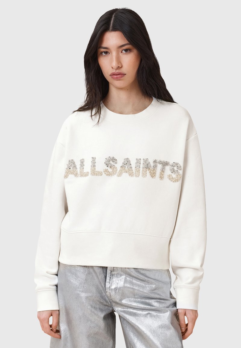 Felpa crema con "ALLSAINTS" in lettere metalliche testurizzate. Presenta un collo rotondo e una vestibilità rilassata. Abbinata a pantaloni argentati lucidi.