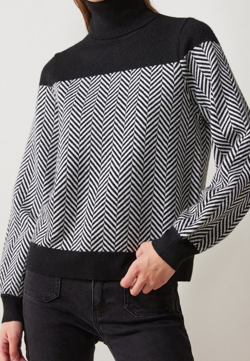 Pull à col roulé en motif chevron noir et blanc avec poignets, ourlet et partie supérieure de la poitrine unis en noir, porté avec un pantalon noir.