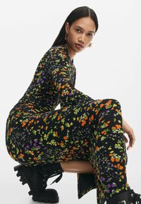 Fekete virágmintás ruha élénk narancssárga, lila és zöld mintákkal, hosszú ujjal, testhez simuló sziluettel, amelyet nehéz fekete bakancsokkal párosítottak.