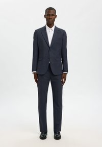 Σκούρο μπλε σακάκι και παντελόνι σετ, σε εφαρμοστή γραμμή, με μία τσέπη στο στήθος και δύο μπροστινές τσέπες. Λευκό πουκάμισο από κάτω, μαύρα παπούτσια.