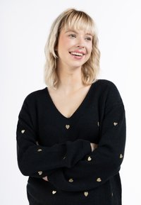 Pulover negru tricotat cu decolteu în V, cu mici modele de inimioare aurii. Textură moale, cu mâneci lungi și croială relaxată.