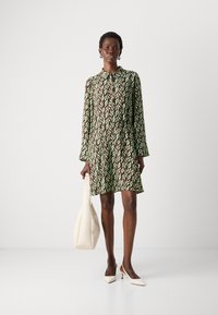 Abito verde e marrone a fantasia con colletto e maniche lunghe, caratterizzato da una gonna svasata. Abbinato a una borsa color crema e scarpe bianche a punta.