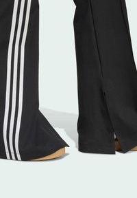 Black athletic pants with three white vertical stripes along the sides. Flared bottom with slits. Smooth fabric and a contrasting light brown sole.