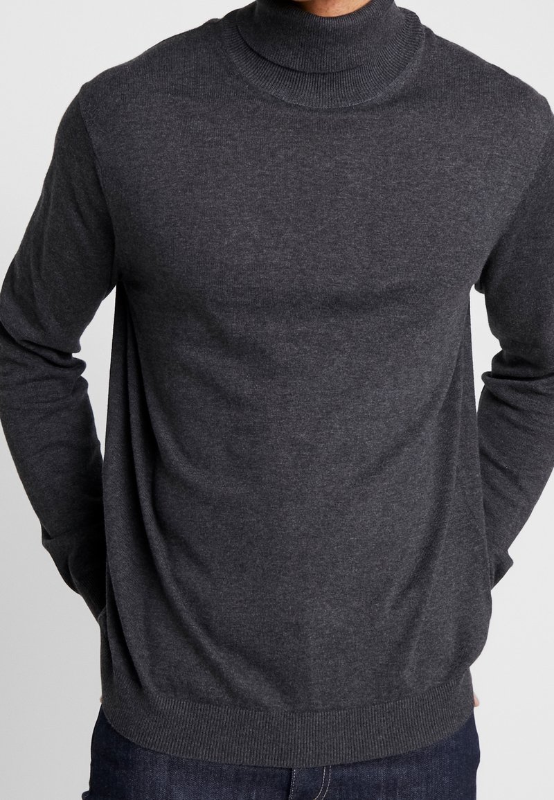 Man in een donkergrijze lange mouwen coltrui met zijn handen casual in de zakken, gecombineerd met donkerblauwe jeans.