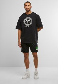 Mann steht mit Händen hinter dem Rücken, trägt ein schwarzes Colucci-T-Shirt, schwarze Shorts mit grün-gelbem Patch, weiße Socken und weiße Turnschuhe.