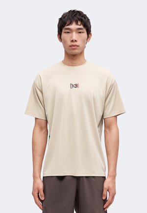 Jeune homme aux cheveux courts et foncés portant un t-shirt beige avec un petit motif noir et rouge centré sur la poitrine et un pantalon foncé.
