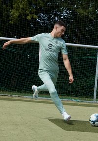 Lichtgroene voetbaloutfit met roze accenten, aansluitend ontwerp en logo zichtbaar. Speler schopt een voetbal op een grasachtige ondergrond.