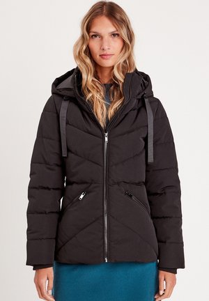 Donna con lunghi capelli biondi ondulati che indossa una giacca imbottita nera con cappuccio e tasche con cerniera, in piedi contro uno sfondo chiaro semplice.