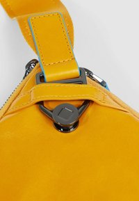 Gele leren band met een gladde textuur en metalen beslag. Heeft een blauwe accentkleur en een glanzende afwerking bij het ritssluitinggebied.