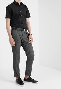 Man med kortärmad svart skjorta med knappar, grå mönstrade byxor, svart bälte, svart klocka och svarta sneakers med vita sulor.