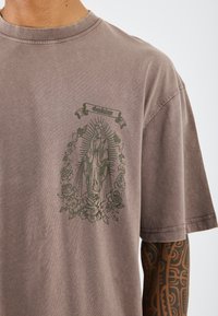 Rudas medvilnės marškinėliai su žalia grafika, vaizduojančia figūrą, apsuptą rožių ir spindulių, su tekstu "Guadalupe" viršuje.
