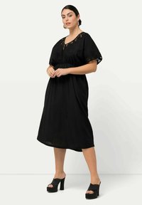 Vestido de algodón negro con un corte holgado, mangas cortas y anchas, detalles de encaje en el escote y longitud hasta la mitad de la pantorrilla, combinado con sandalias de plataforma negras.