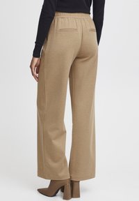 Pantalons larges marron clair avec une taille élastique et deux poches arrière, associés à des talons aiguilles marron. Texture lisse.
