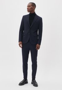 Marineblauer Anzug mit schlanker Passform, Ein-Knopf-Verschluss und zwei vorderen Taschen. Tragen über einem schwarzen Rollkragenpullover und kombiniert mit schwarzen Slippern.