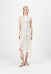 Obleka iz čipke z visokim ovratnikom, brez rokavov, asimetričen rob, v off-white barvi, plastnata tekstura. Model nosi zaponkaste bež petke.