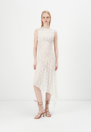 Lace dress with a high neckline, sleeveless design, asymmetrical hem, in off-white, layered texture. Model wears strappy beige heels.