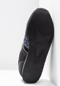 Semelle de chaussure noire avec motif de crampons en forme d'étoile et "VERSACE JEANS" embossé au centre, présenté sur fond blanc.